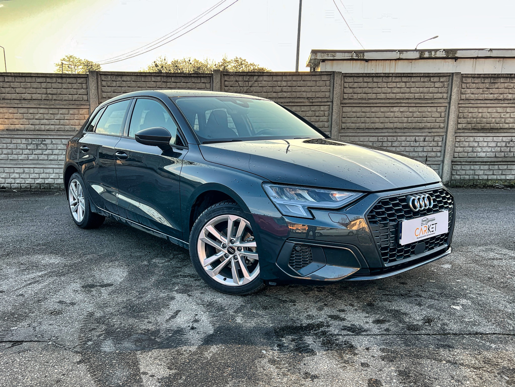 Ciemny hatchback Audi A3 z tabliczką firmy na tle betonowego muru.