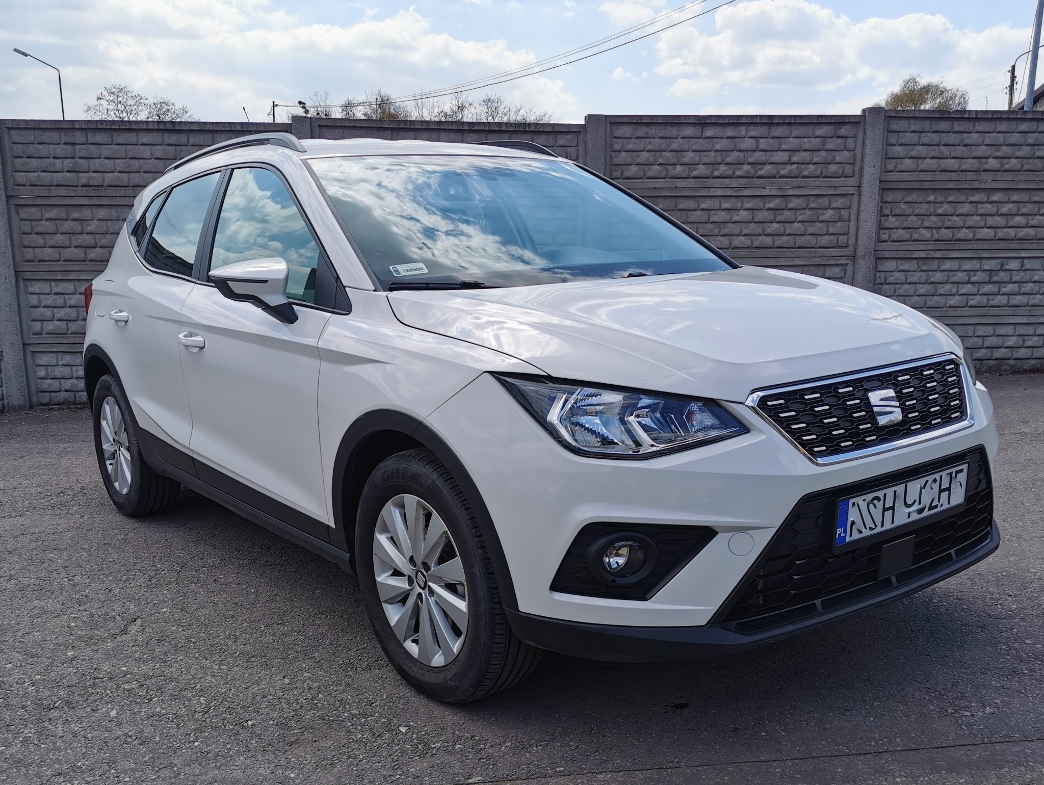 Biały crossover Seat Arona zaparkowany na zewnątrz, widok z przodu, z widoczną tablicą rejestracyjną i odbiciem chmur w szybie.