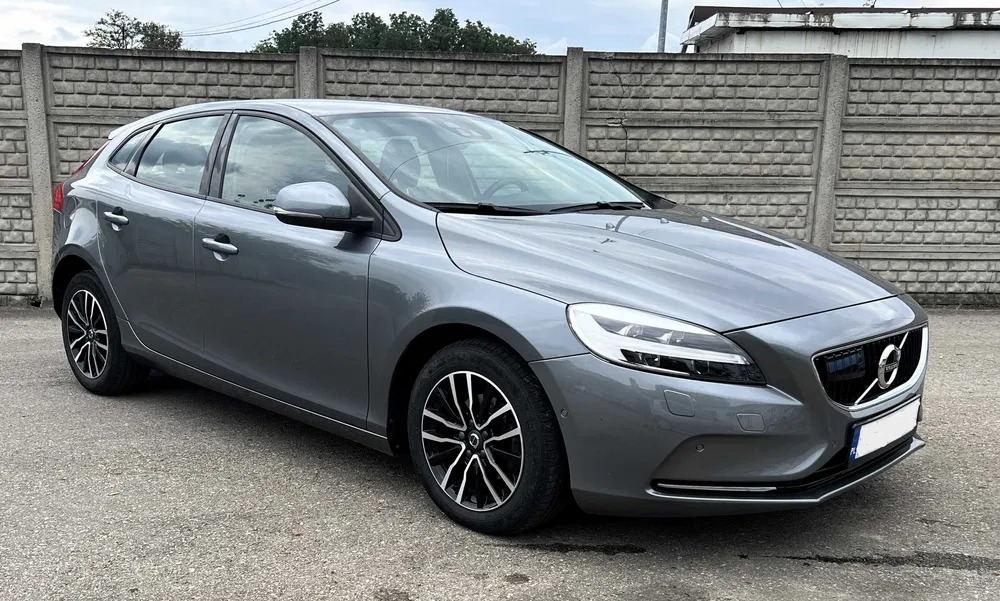 Szary hatchback Volvo V40 z czarno-srebrnymi felgami, zaparkowany na tle betonowego muru z widocznymi śladami użytkowania.