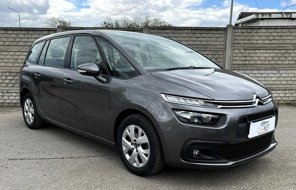 Szary minivan Citroen z logo firmy na przednim zderzaku, zaparkowany na zewnątrz z szarym ceglanym murem w tle.