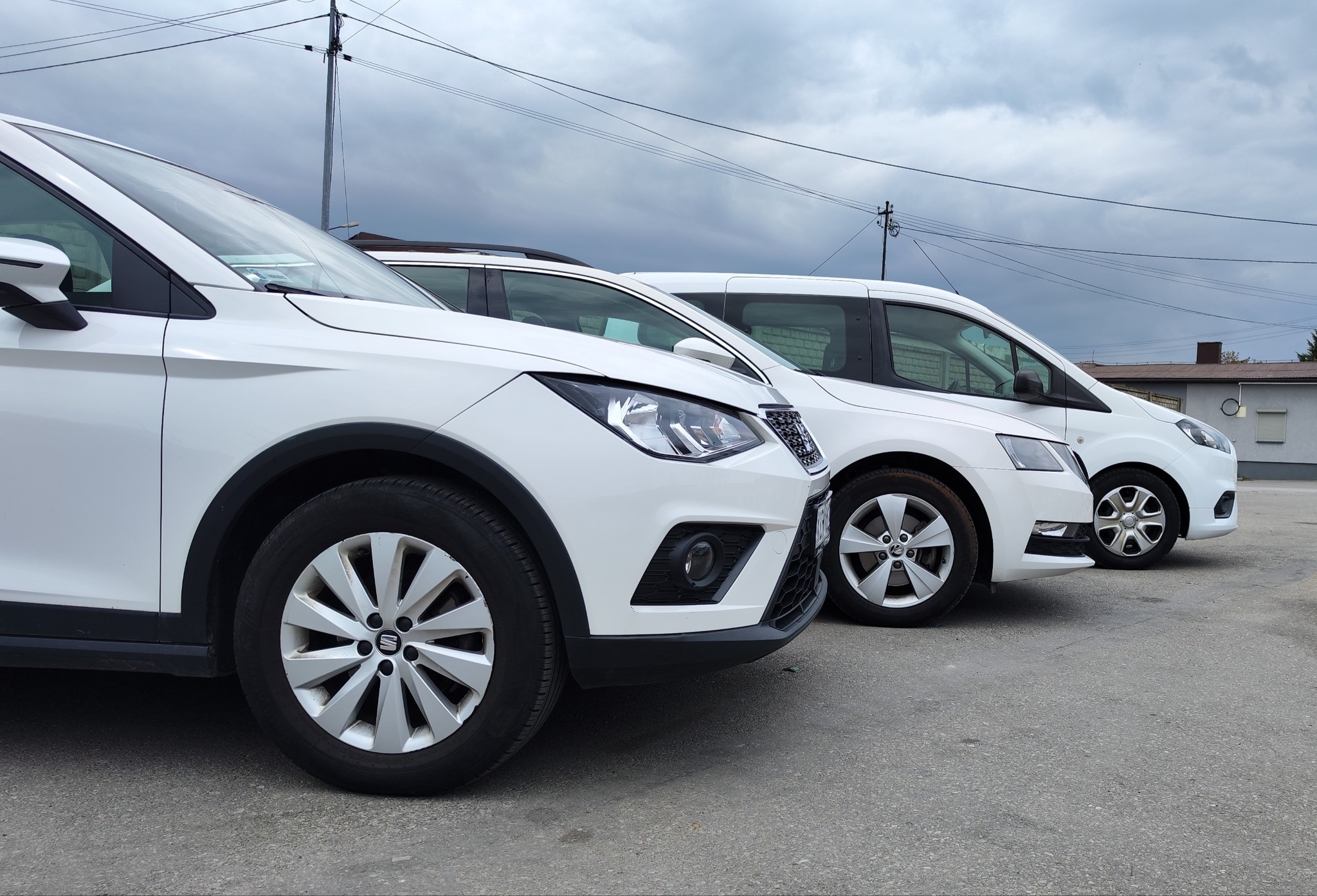 Rząd białych samochodów osobowych i dostawczych na parkingu, widok z boku, pochmurne niebo w tle.