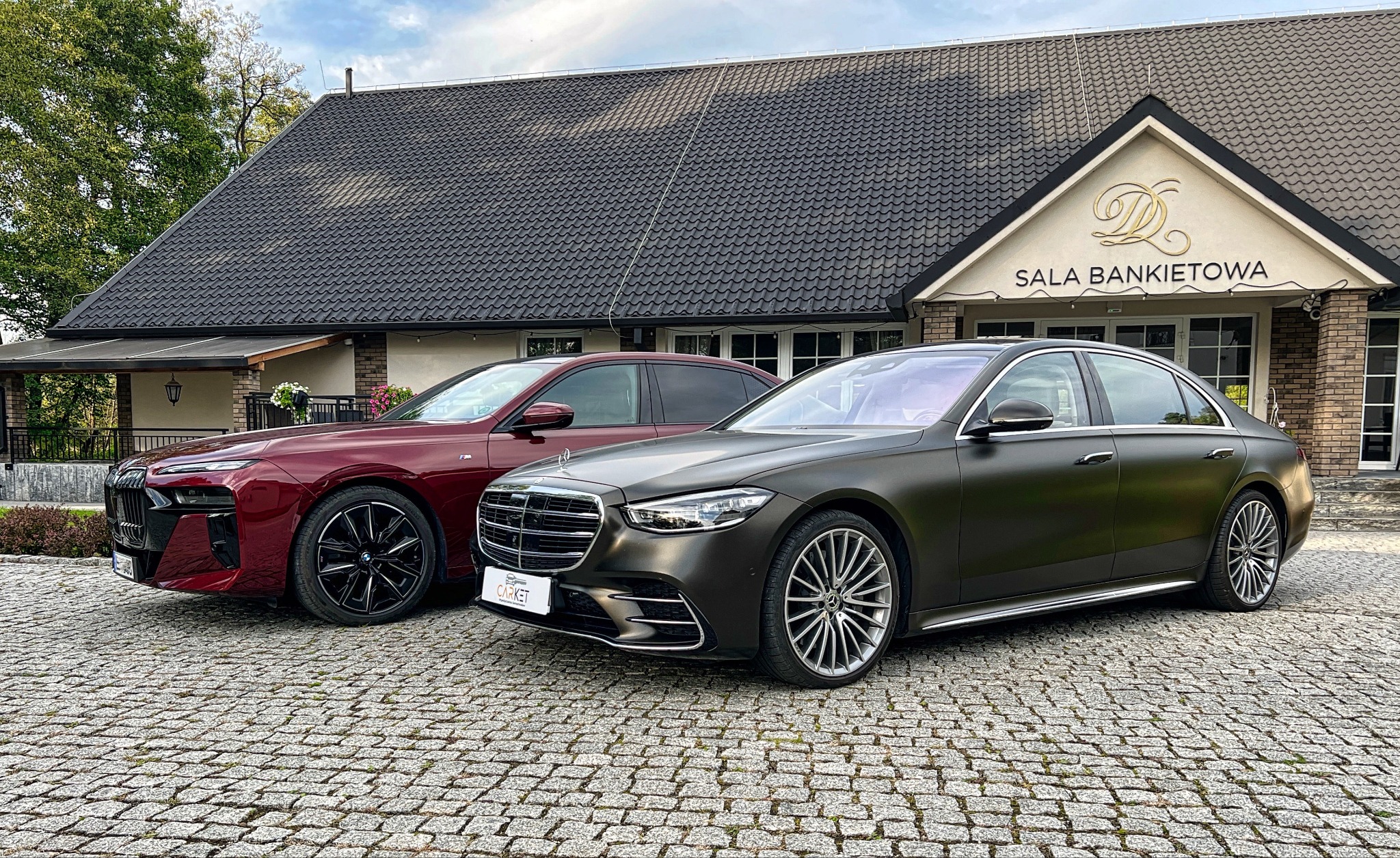 Dwa luksusowe samochody: bordowy BMW i7 i matowy, grafitowy Mercedes-Benz Klasy S, zaparkowane przed salą bankietową z brukowanym podjazdem.