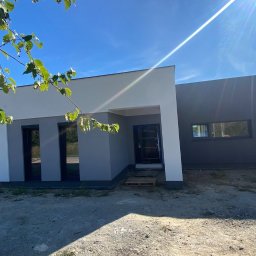 happyhouse-developments - Nowoczesny dom murowany z płaskim dachem, pomalowany na szaro i biało, z widocznymi oknami i drzwiami, otoczony niezagospodarowanym terenem i zielenią.