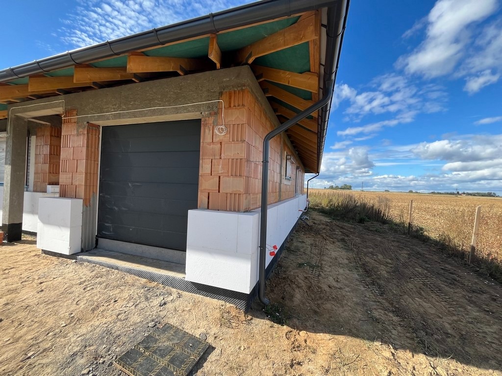 Surowy stan domu z garażem, widoczne bloczki ceramiczne, ocieplenie styropianem fundamentów, ciemna brama garażowa, rynna spustowa, w tle pole uprawne pod błękitnym niebem.