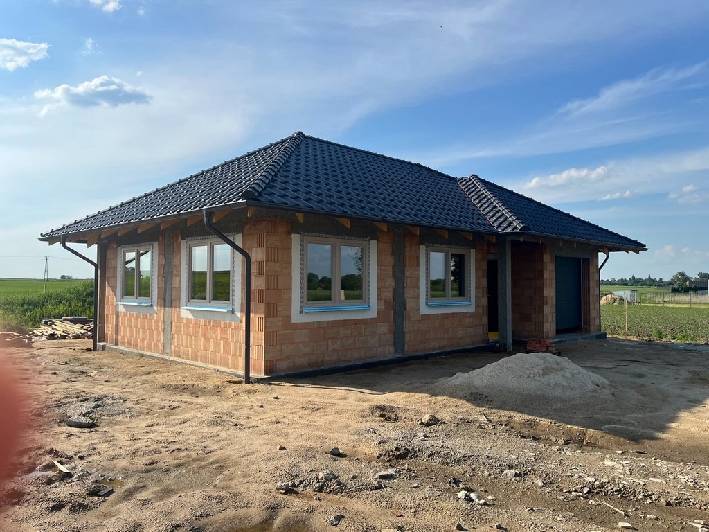 Niski, parterowy budynek w trakcie budowy z czerwonej cegły, z czarnym dachem i białymi ramami okiennymi, widok z zewnątrz na tle zielonego pola i błękitnego nieba.