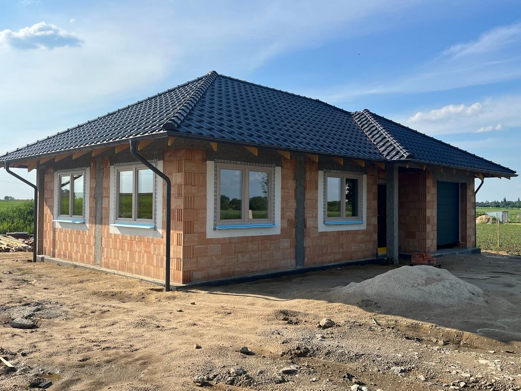 Niedokończony parterowy budynek z czarną dachówką i oknami z jasnymi ramami w trakcie budowy, widoczny stos piasku przed budynkiem.