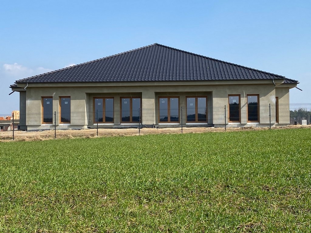 Surowy stan elewacji parterowego domu z ciemną dachówką, widoczne okna z drewnianą stolarką, ogrodzenie z siatki i trawnik w pierwszym planie.