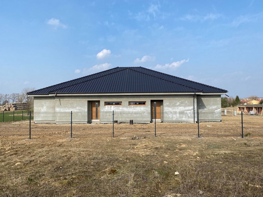 Surowy, parterowy dom jednorodzinny z ciemną dachówką, ogrodzony siatką, w trakcie budowy, widoczne ślady po tynku na elewacji, z dwoma brązowymi drzwiami wejściowymi i małymi oknami.