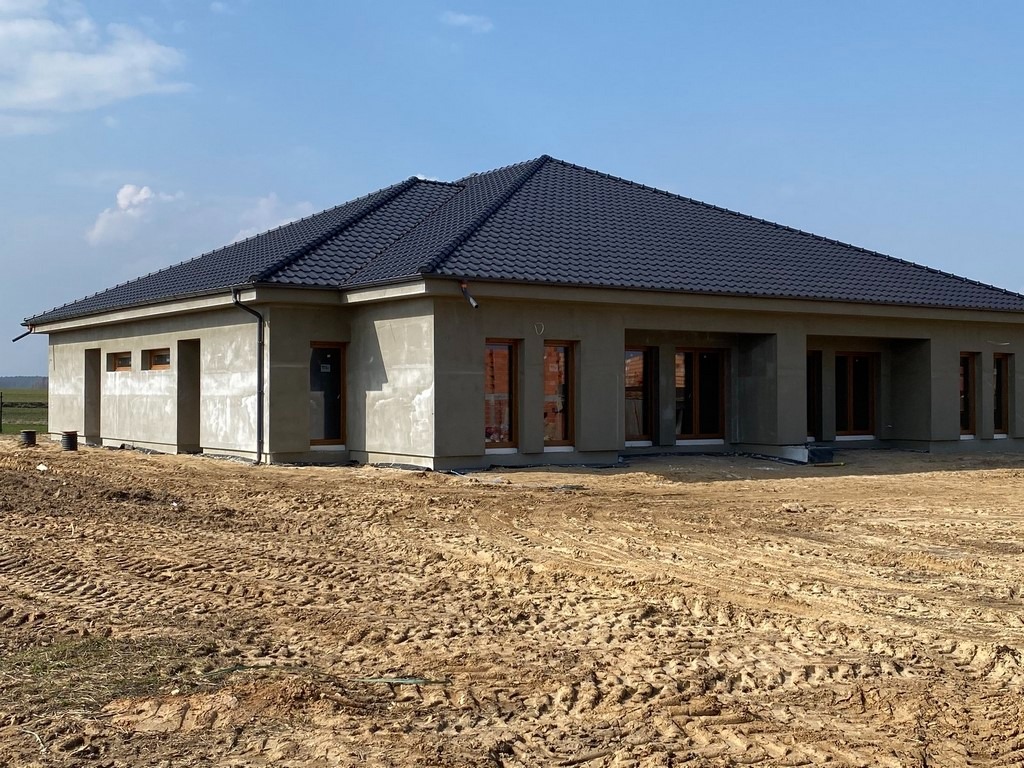 Niedokończony parterowy dom z czarną dachówką i surowymi ścianami zewnętrznymi, widoczne drewniane ramy okienne, teren wokół domu z wyraźnymi śladami opon.