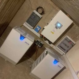 Elektrowniesloneczne.tech - Kompletna instalacja magazynu energii Kon-Tec w surowym pomieszczeniu z betonowymi ścianami. Widoczne dwa akumulatory, inwerter i rozdzielnica.