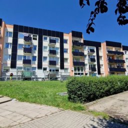 Elektrowniesloneczne.tech - Blok z panelami fotowoltaicznymi na balkonach, widoczny z poziomu trawnika. Na pierwszym planie kobieta i zaparkowany biały bus. Montaż paneli w toku.