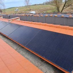 Elektrowniesloneczne.tech - Montaż paneli fotowoltaicznych na dachu z pomarańczowej dachówki. Widoczne panele w rzędzie, zabezpieczenia i krajobraz w tle. Rękawice robocze na dachu.