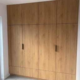 LAJTNER INTERIORS SPÓŁKA Z OGRANICZONĄ ODPOWIEDZIALNOŚCIĄ - Pełna zabudowa ścienna z szaf z drewnopodobnymi frontami, czarne, minimalistyczne uchwyty, szara podłoga, oświetlenie sufitowe.