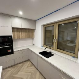 LAJTNER INTERIORS SPÓŁKA Z OGRANICZONĄ ODPOWIEDZIALNOŚCIĄ - Jasnoszara kuchnia na wymiar z drewnianymi akcentami i czarnym zlewem pod oknem, parkiet w jodełkę na podłodze.