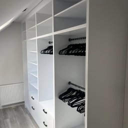LAJTNER INTERIORS SPÓŁKA Z OGRANICZONĄ ODPOWIEDZIALNOŚCIĄ - Biała, zabudowana szafa z półkami i szufladami, czarne wieszaki na ubrania, poddasze ze skosem, szara podłoga laminowana.