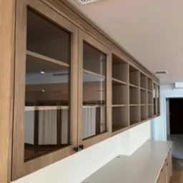 Długa, zabudowana szafka wisząca z przeszklonymi drzwiczkami i otwartymi półkami, wykonana z jasnego drewna, z widocznym systemem wentylacji sufitowej