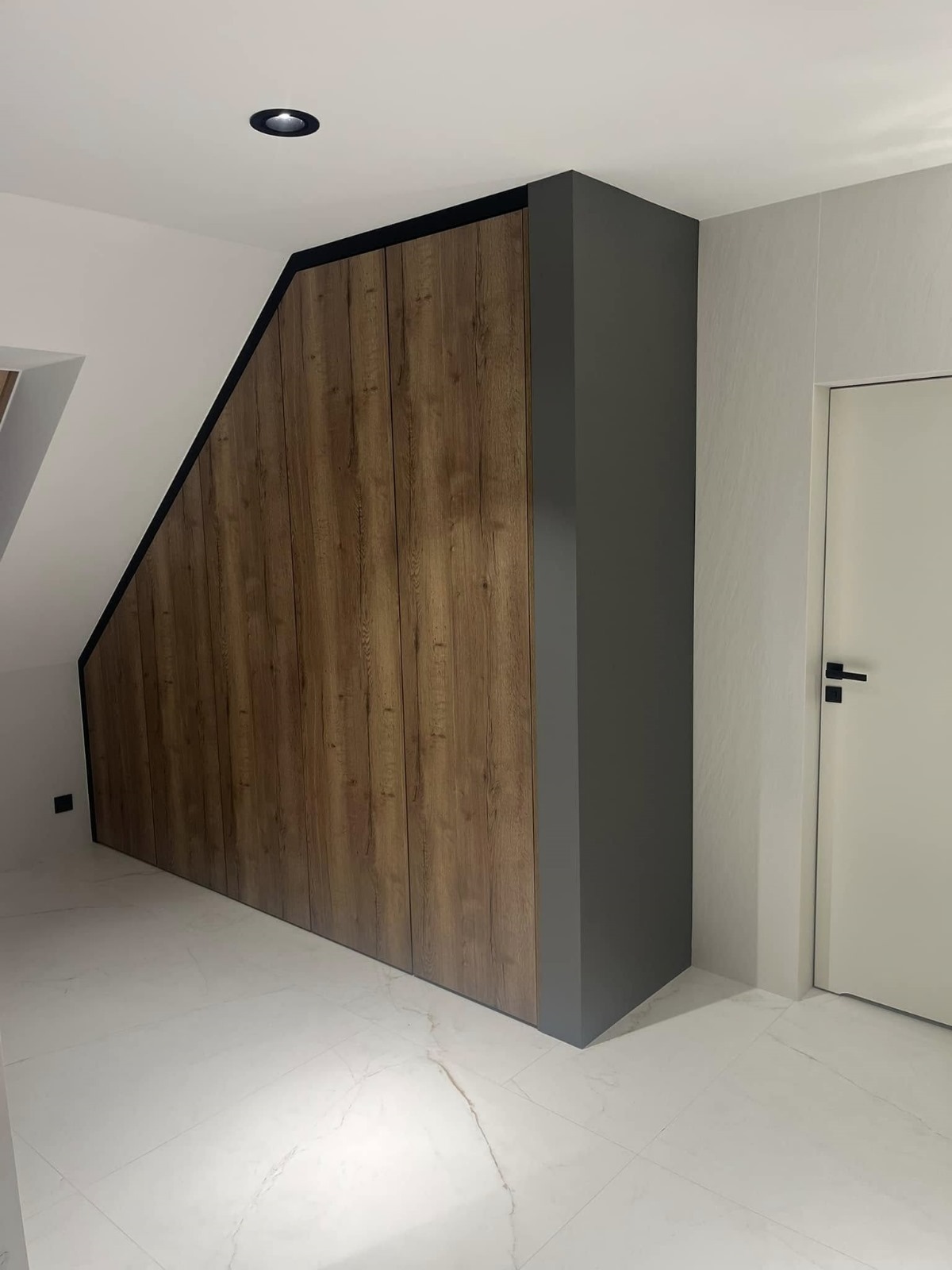 Szafa wnękowa z drewnianymi frontami, dopasowana do skosu poddasza, z szarą obudową, na białej marmurowej podłodze, oświetlenie punktowe w suficie.