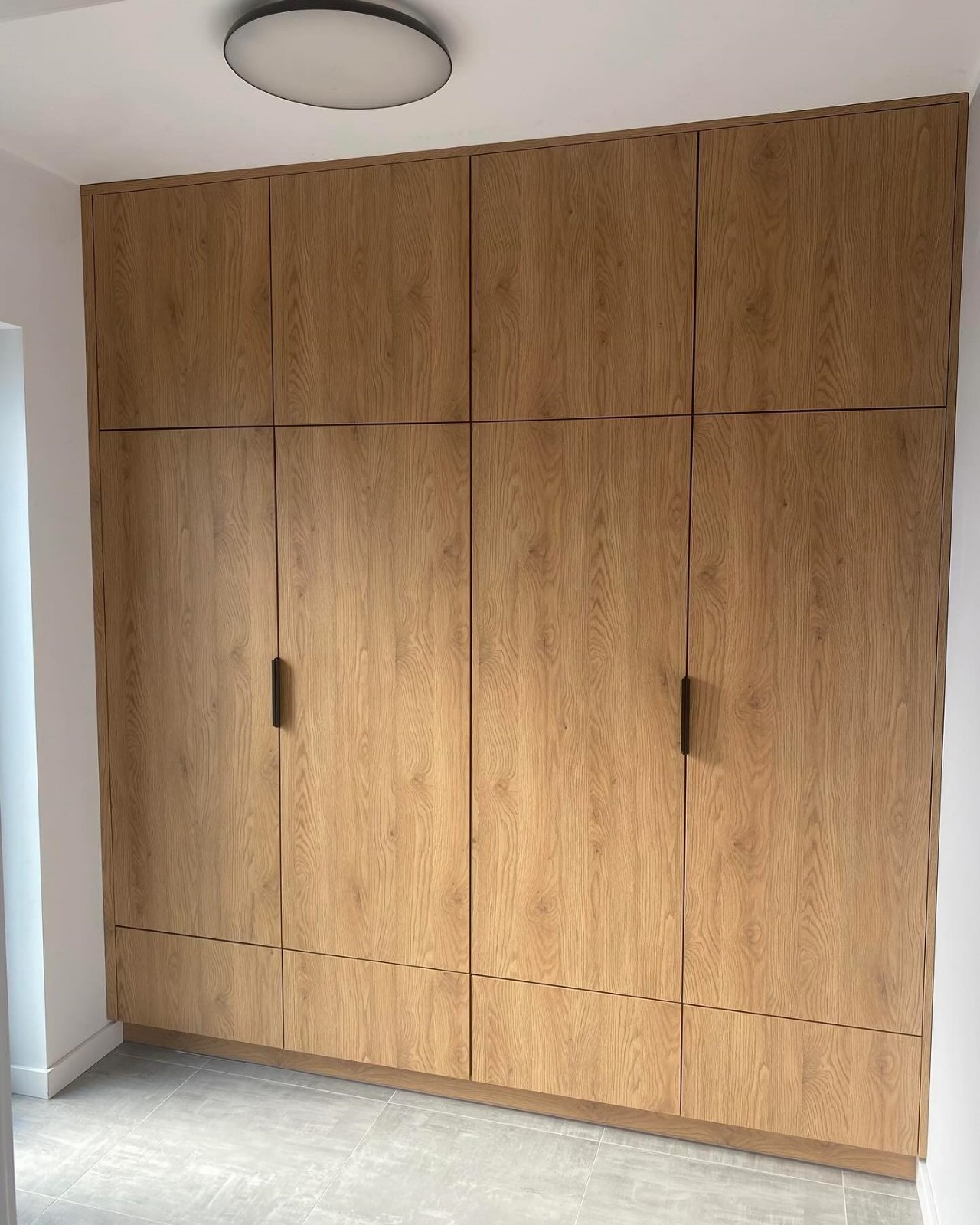 Pełna zabudowa ścienna z szaf z drewnopodobnymi frontami, czarne, minimalistyczne uchwyty, szara podłoga, oświetlenie sufitowe.