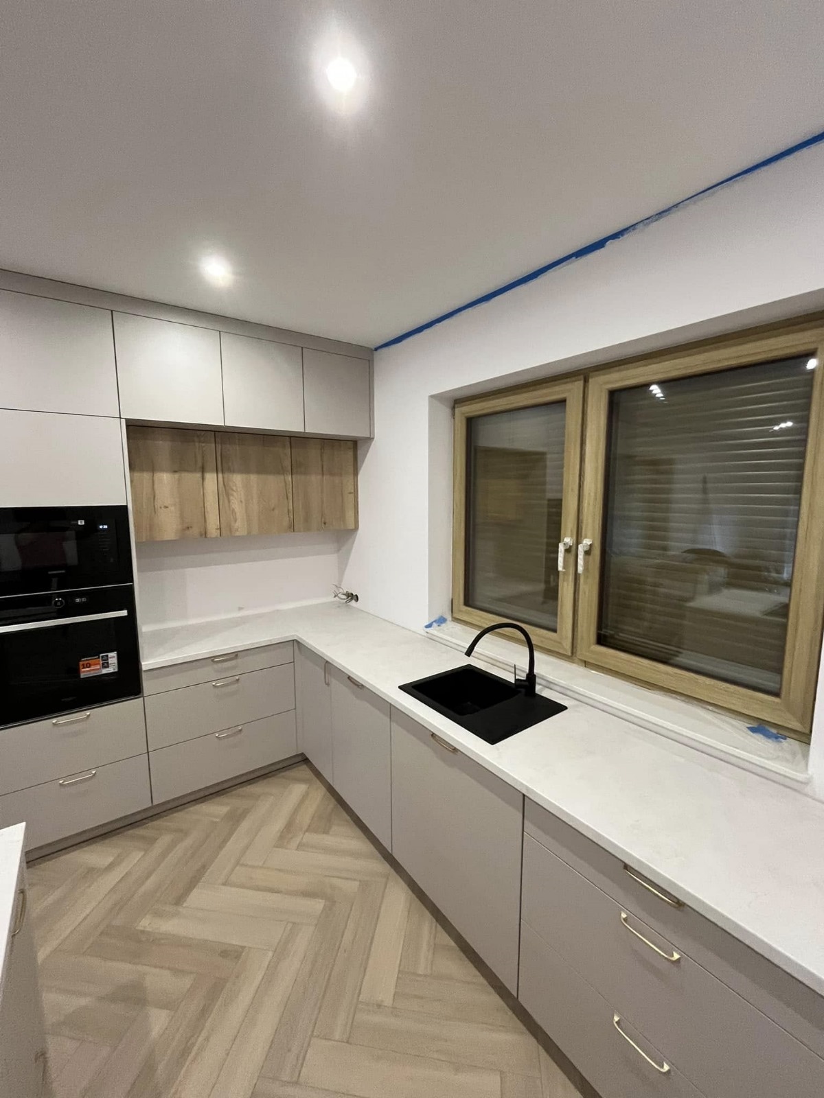 Jasnoszara kuchnia na wymiar z drewnianymi akcentami i czarnym zlewem pod oknem, parkiet w jodełkę na podłodze.