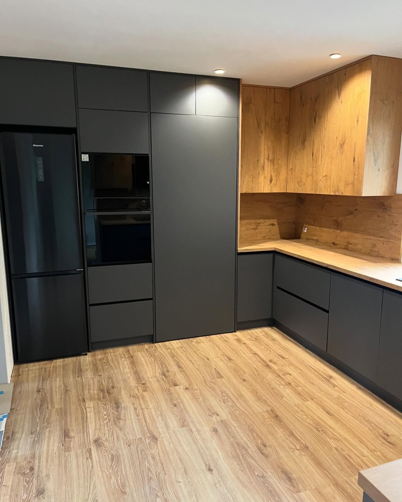 Nowoczesna kuchnia z grafitowymi frontami, drewnopodobnym blatem i fornirem na ścianie, zintegrowana zabudowa z lodówką i piekarnikiem.