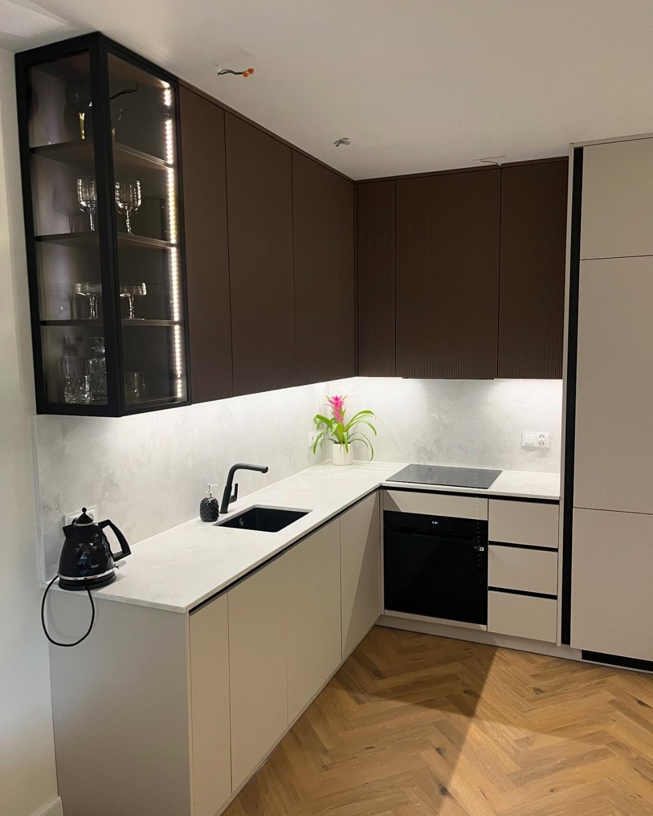 Nowoczesna kuchnia z brązowymi ryflowanymi szafkami górnymi, białymi szafkami dolnymi, czarną armaturą, szklaną witryną z podświetleniem LED i drewnianą podłogą w jodełkę.