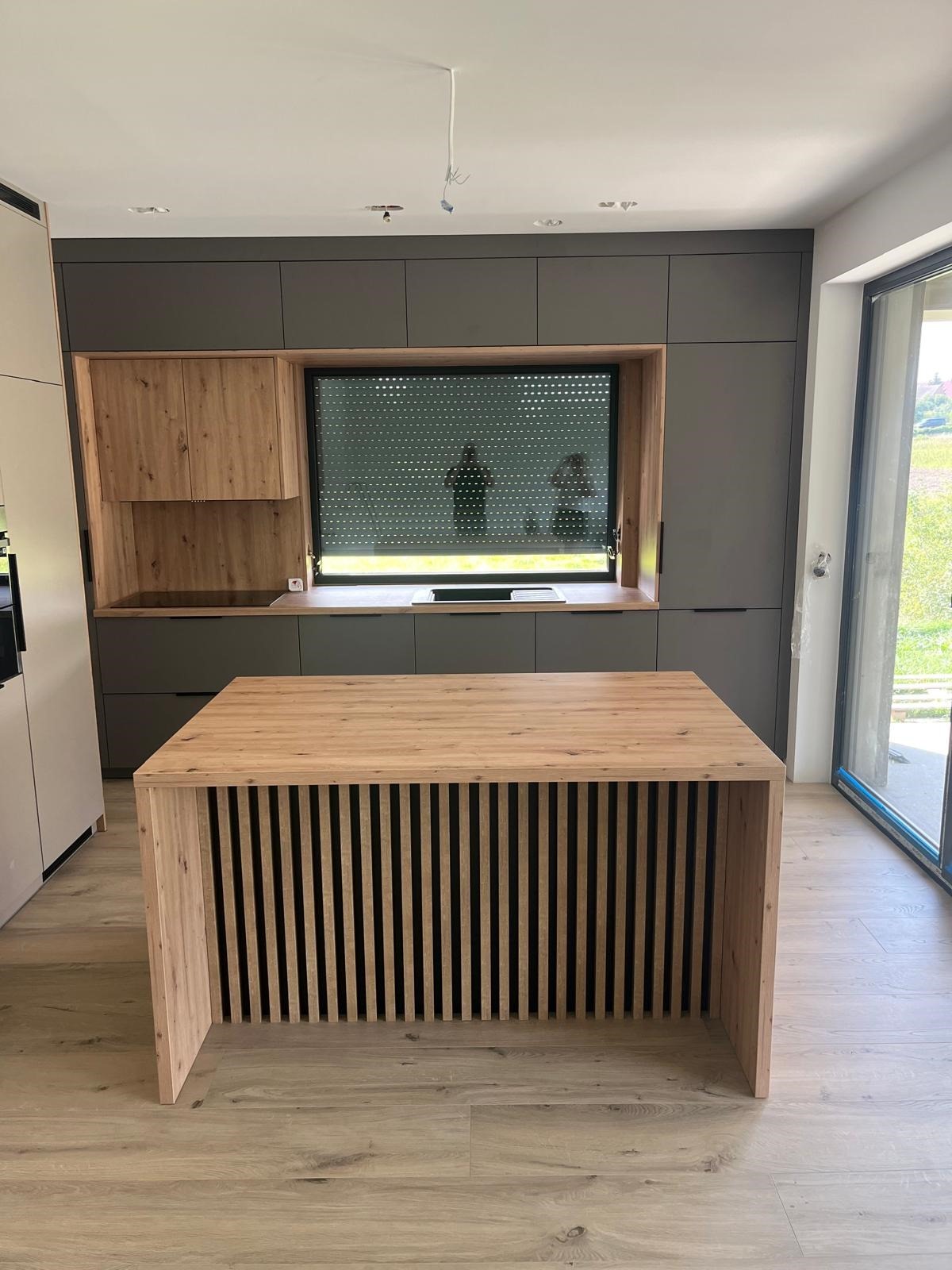 Nowoczesna kuchnia z wyspą o drewnianym wykończeniu, szare szafki wiszące i okno z żaluzjami zewnętrznymi.