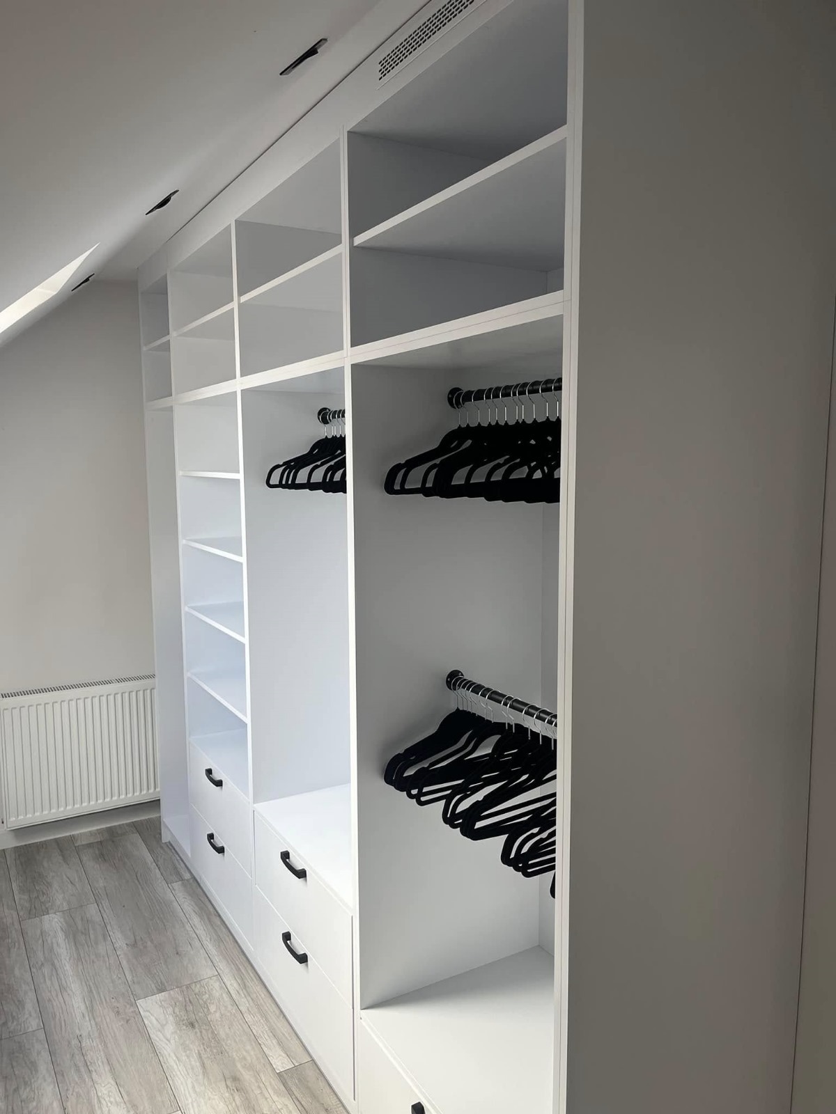 Biała, zabudowana szafa z półkami i szufladami, czarne wieszaki na ubrania, poddasze ze skosem, szara podłoga laminowana.