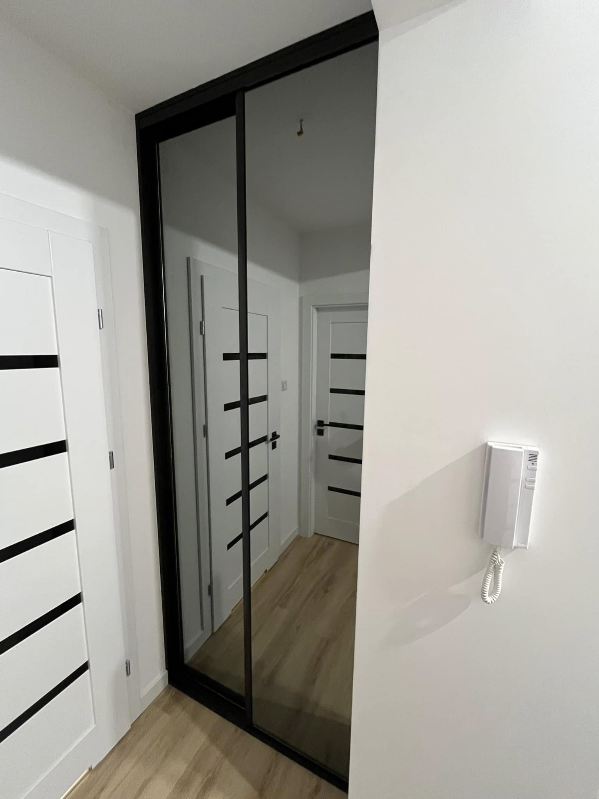 Szafa wnękowa z przesuwnymi drzwiami lustrzanymi w czarnej ramie, odbijająca białe drzwi z czarnymi poziomymi paskami w korytarzu z panelami na podłodze, na ścianie domofon.