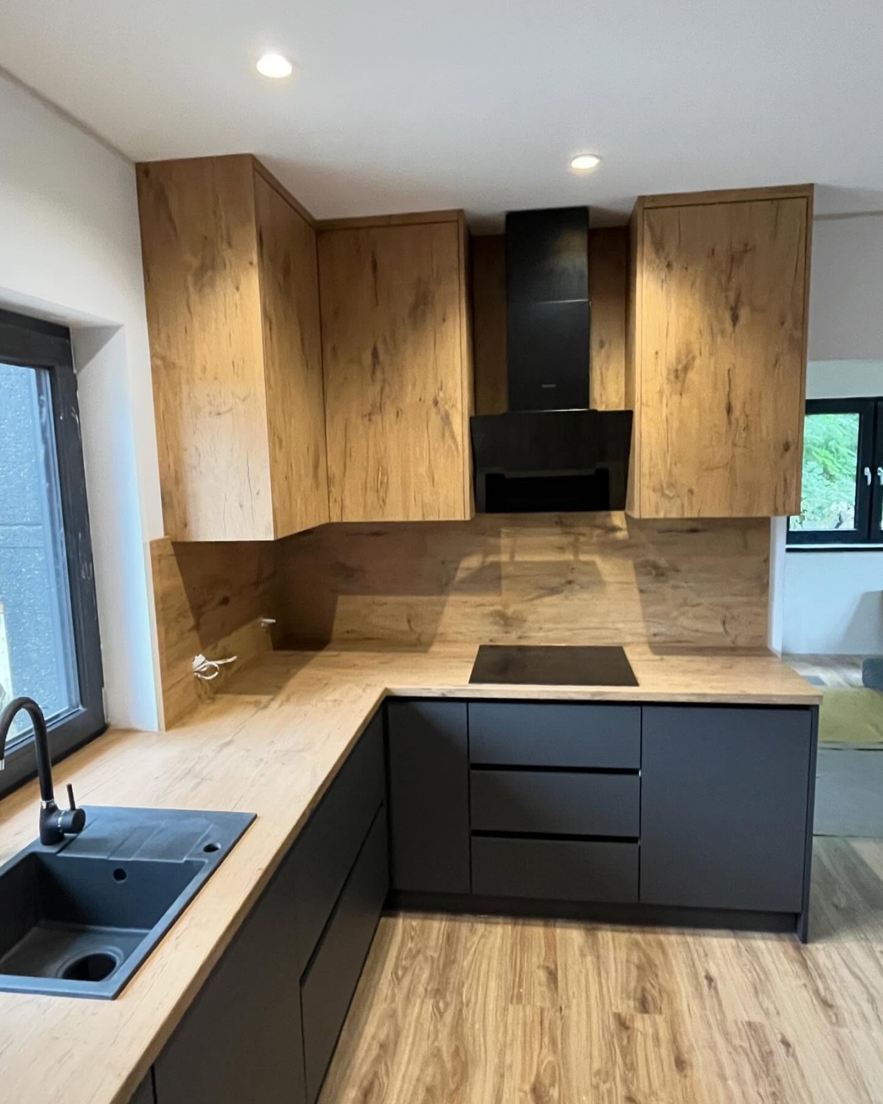 Nowoczesna kuchnia z drewnianymi szafkami górnymi i czarnymi szafkami dolnymi, czarny okap nad płytą grzewczą, drewniany blat i podłoga.