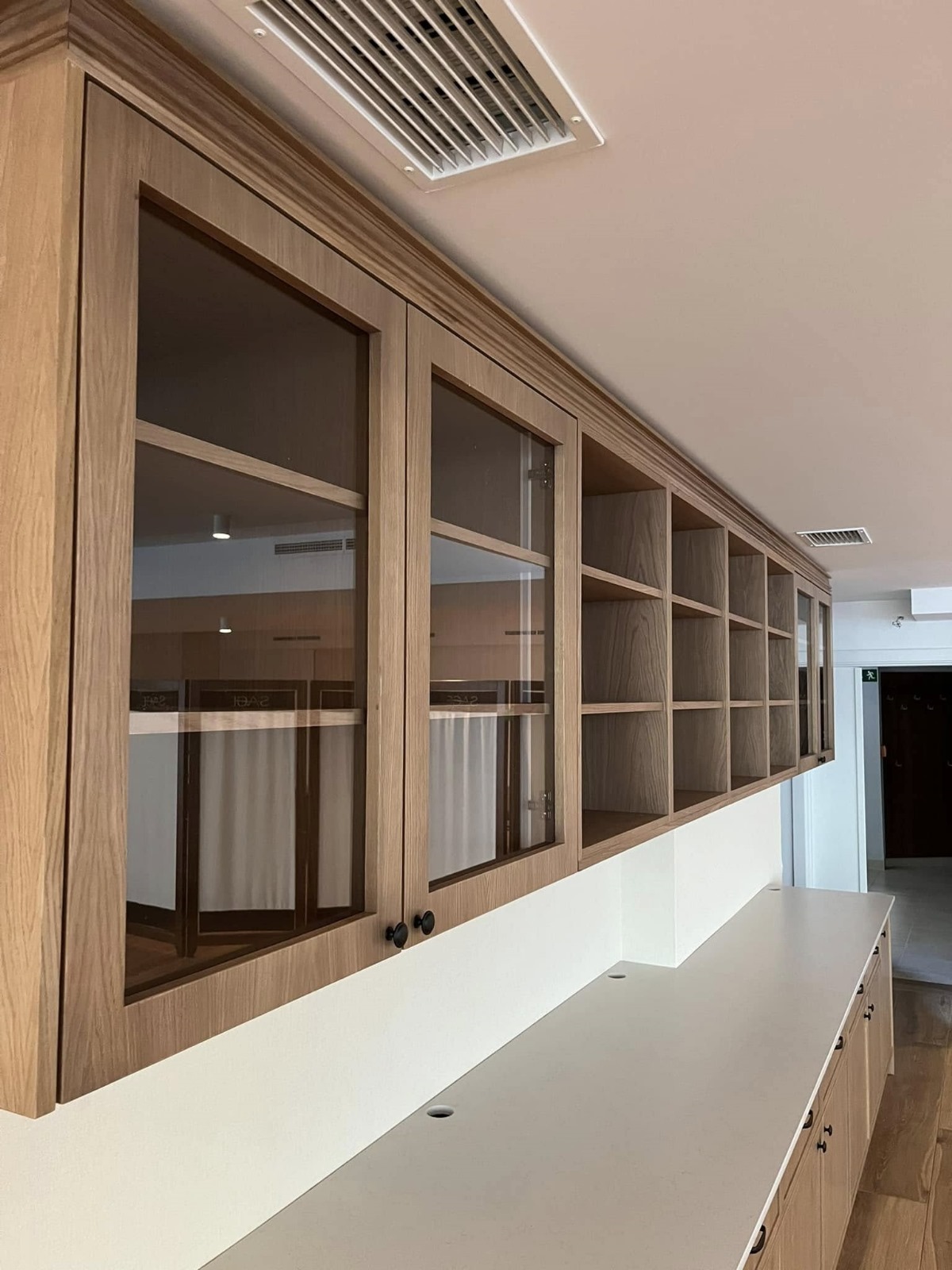 Długa, zabudowana szafka wisząca z przeszklonymi drzwiczkami i otwartymi półkami, wykonana z jasnego drewna, z widocznym systemem wentylacji sufitowej