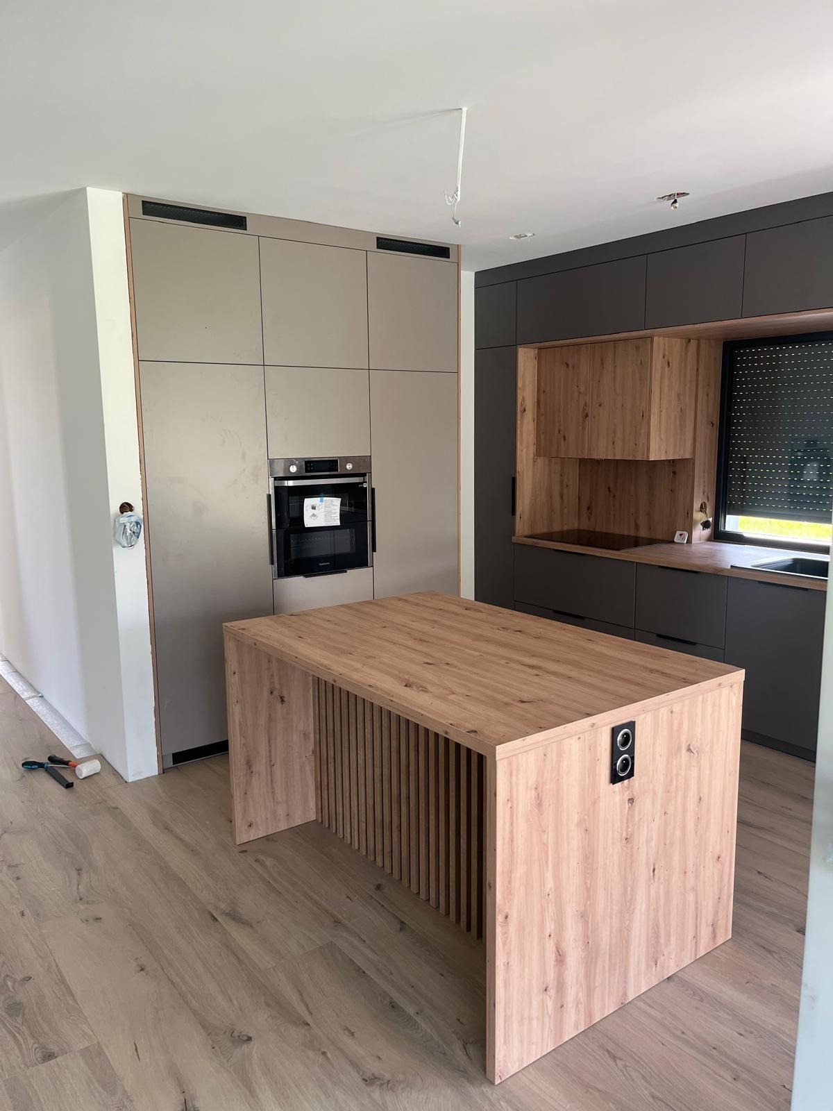 Nowoczesna kuchnia z wyspą z drewnianym blatem i frontami, beżowe i grafitowe szafki bez uchwytów, wbudowany piekarnik i płyta grzewcza, widoczne narzędzia na podłodze.