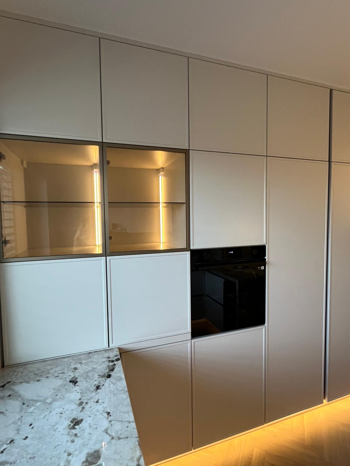 Nowoczesna kuchnia z zabudową sięgającą sufitu, jasne fronty, szklane witryny z oświetleniem LED oraz wbudowany piekarnik.