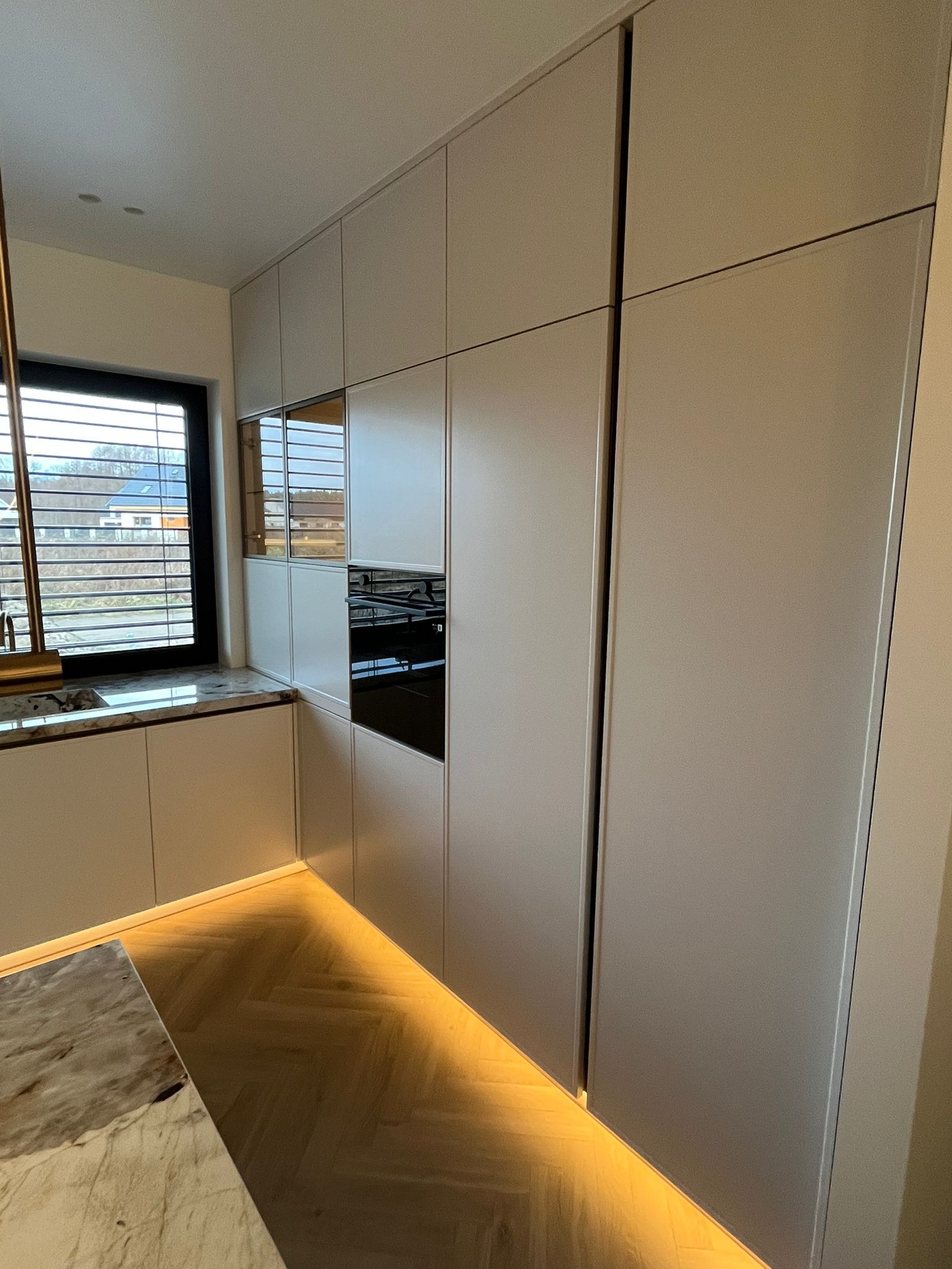 Nowoczesna kuchnia z zabudową sięgającą sufitu, jasne fronty szafek, oświetlenie LED pod szafkami, parkiet w jodełkę, marmurowy blat.