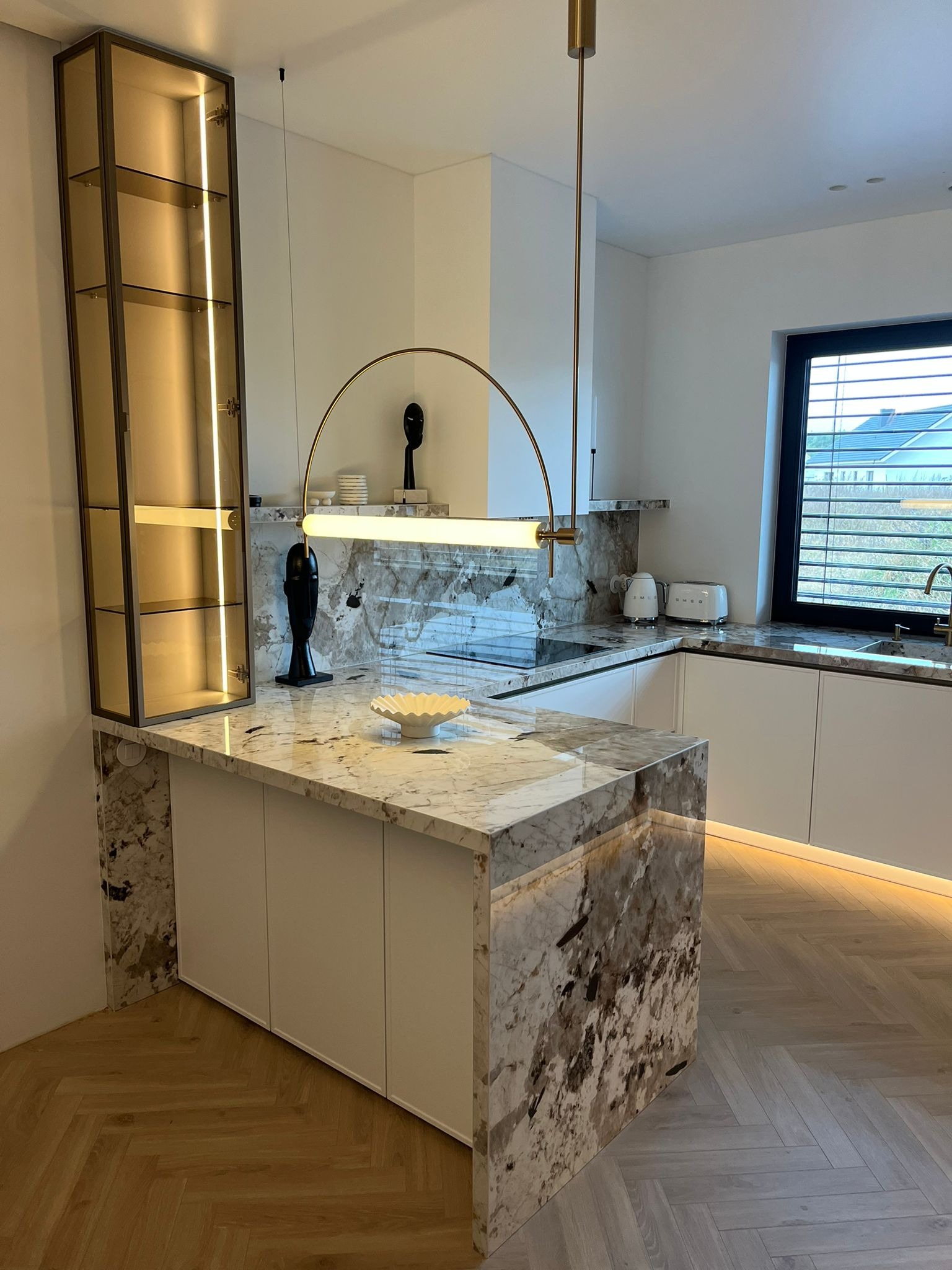 Nowoczesna kuchnia z wyspą wykończoną marmurem, oświetleniem LED pod szafkami i wiszącą lampą łukową w kolorze złotym, na blacie rzeźba i misa dekoracyjna.