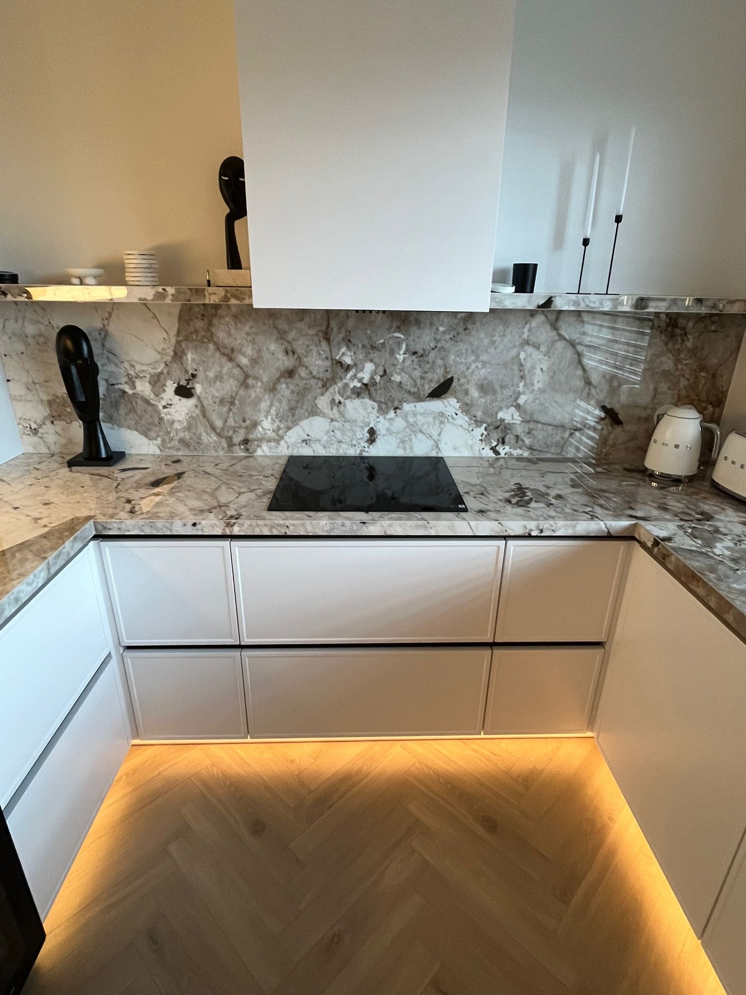 Nowoczesna kuchnia z białymi szafkami, marmurowym blatem i podświetleniem LED pod szafkami dolnymi, widoczna płyta indukcyjna i dekoracyjne figurki.