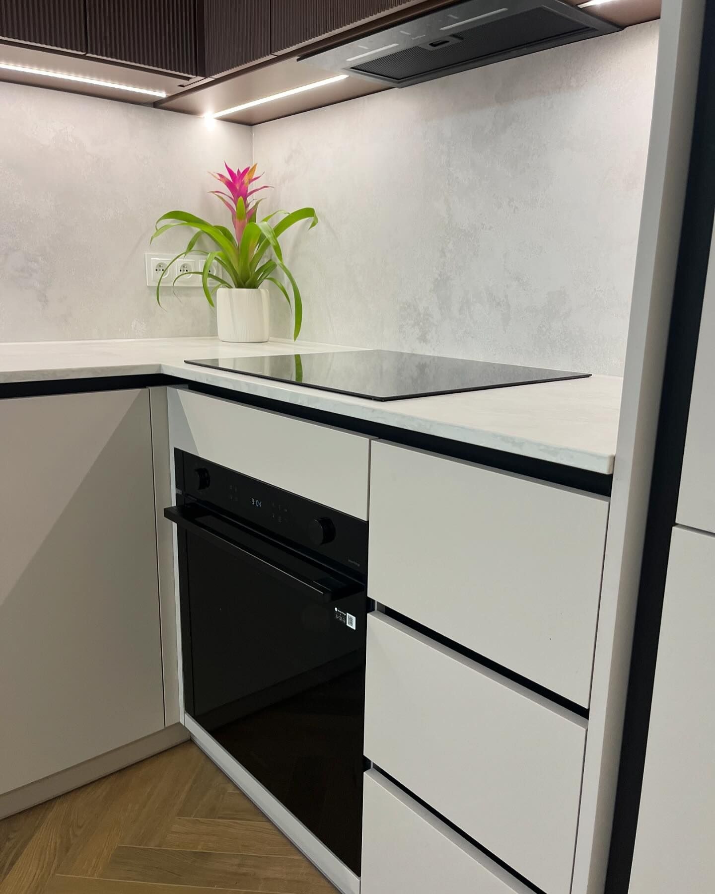 Fragment nowoczesnej kuchni z białymi frontami szafek, czarnym blatem i płytą indukcyjną, na blacie doniczka z różowym kwiatem, widoczny piekarnik i drewniana podłoga w jodełkę.