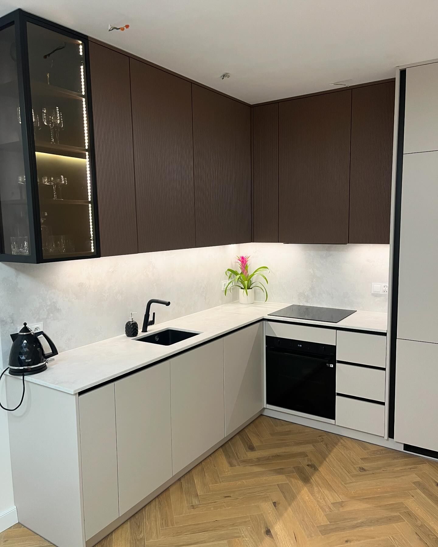 Nowoczesna kuchnia narożna z brązowymi, ryflowanymi szafkami górnymi i jasnymi dolnymi, blatem z jasnego kamienia, czarną armaturą i podświetlaną witryną z kieliszkami.