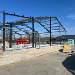 Stalowa konstrukcja hali w trakcie budowy, widoczne przygotowane płyty warstwowe, podnośnik nożycowy i fundamenty.