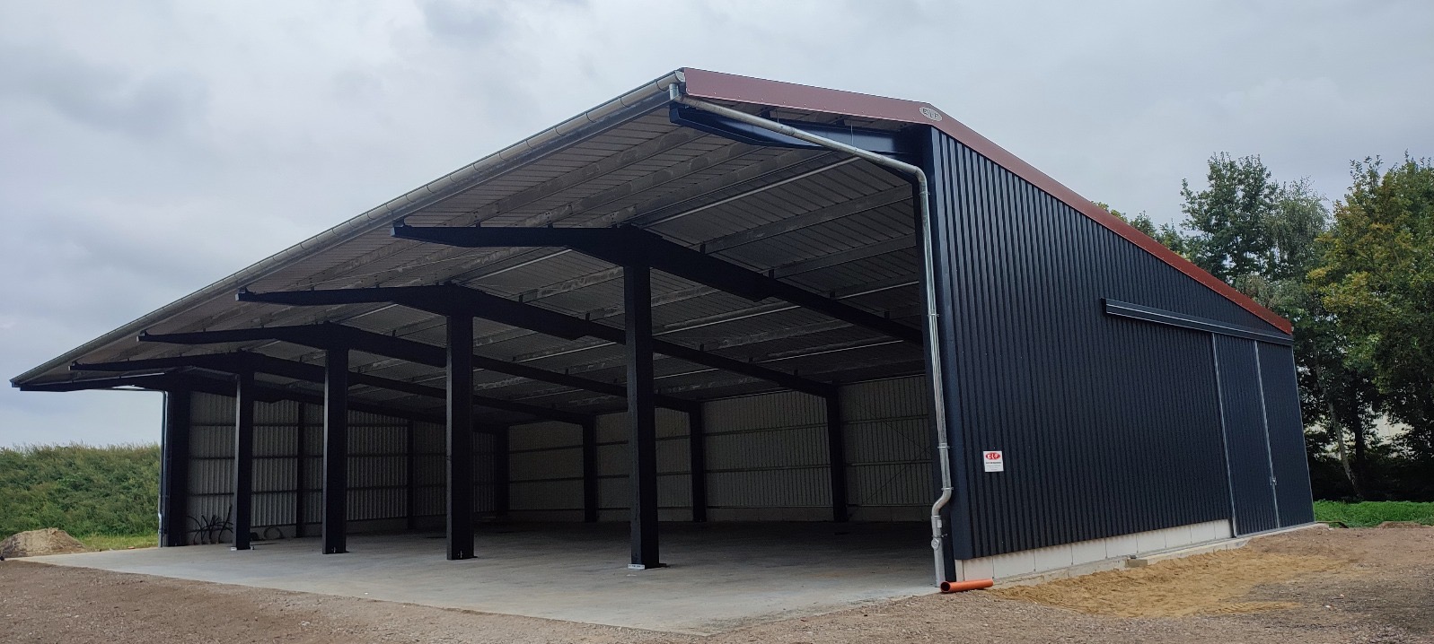 Zakończona budowa otwartej hali magazynowej z płyt warstwowych w Ostrowie Wielkopolskim, widoczne czarne stalowe słupy konstrukcyjne i szara blacha trapezowa na dachu.