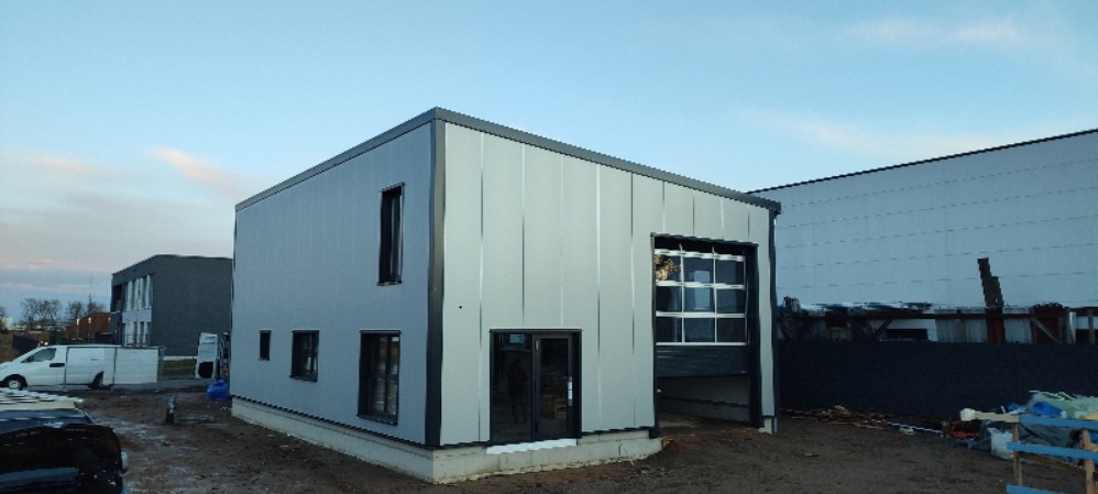 Nowoczesny budynek przemysłowy wykończony płytami warstwowymi, z widocznymi oknami, drzwiami i częściowo otwartą bramą garażową, w tle inne budynki i pojazdy.