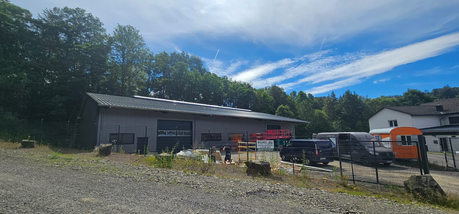 Budynek przemysłowy z ciemnym dachem blaszanym, widoczne prace przygotowawcze i materiały budowlane, w tle zieleń i błękitne niebo z chmurami.