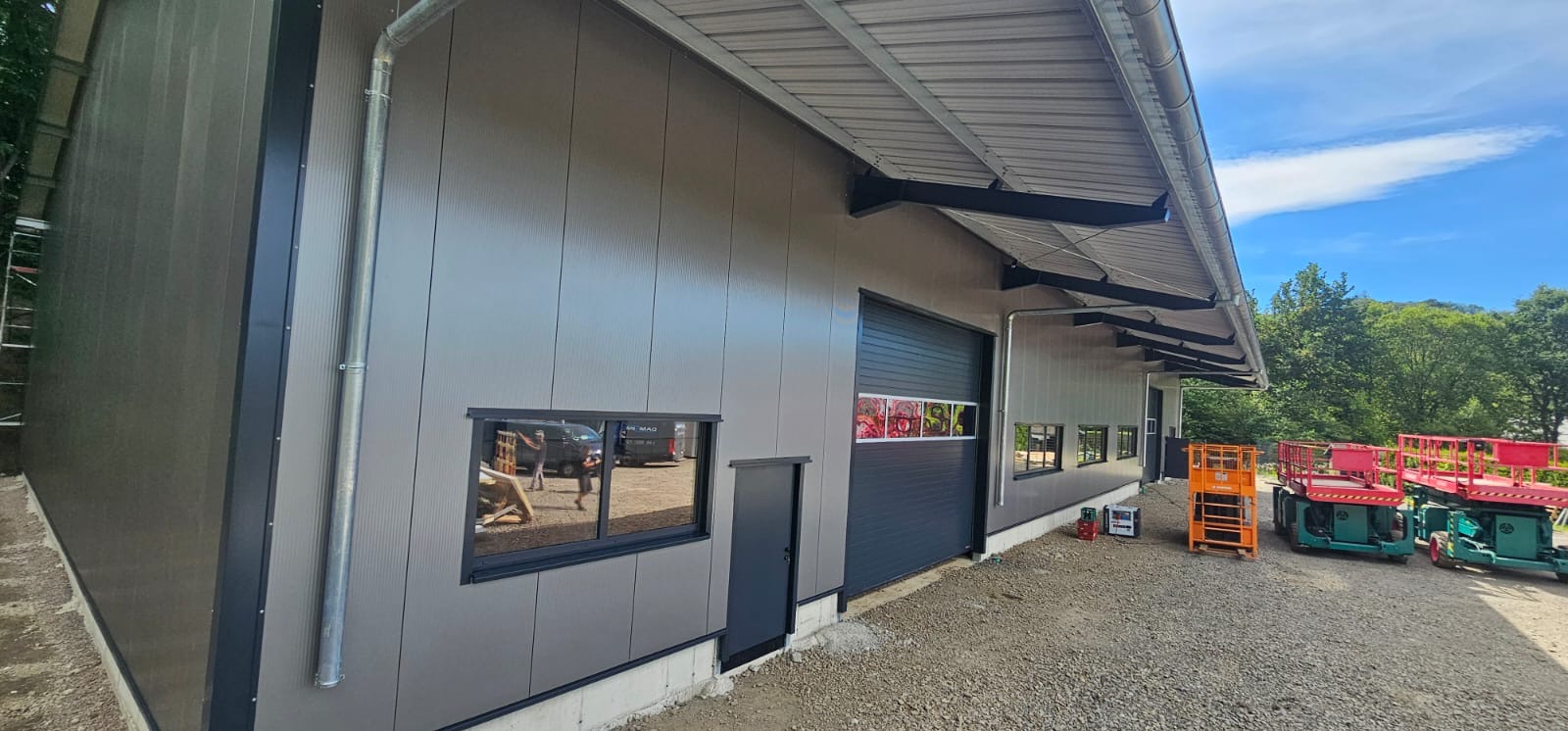 Nowoczesny budynek przemysłowy obłożony szarymi płytami warstwowymi, widoczne okna, brama garażowa, rynna spustowa, oraz podnośniki koszowe na żwirowym podłożu.