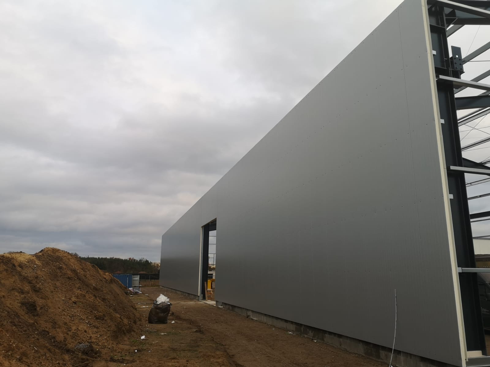 Szara ściana hali przemysłowej z paneli warstwowych w trakcie montażu, widoczna stalowa konstrukcja nośna i sterta ziemi obok.