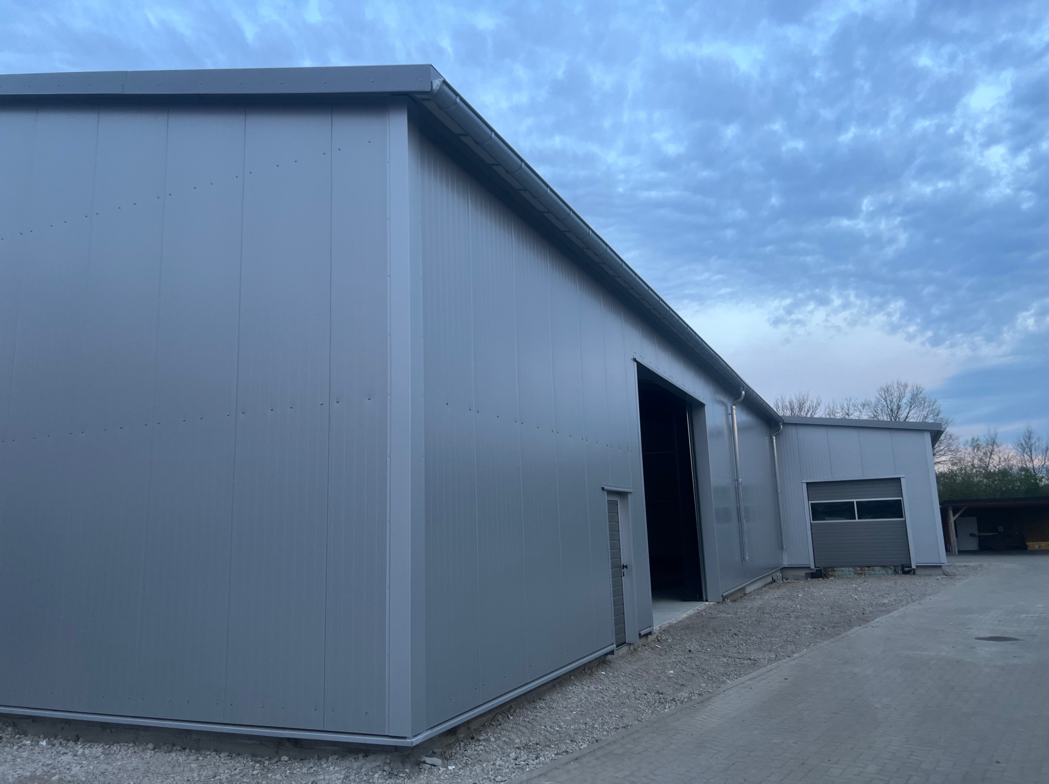 Nowoczesny budynek przemysłowy z szarych płyt warstwowych, widoczne łączenia paneli i brama garażowa, pochmurne niebo w tle.