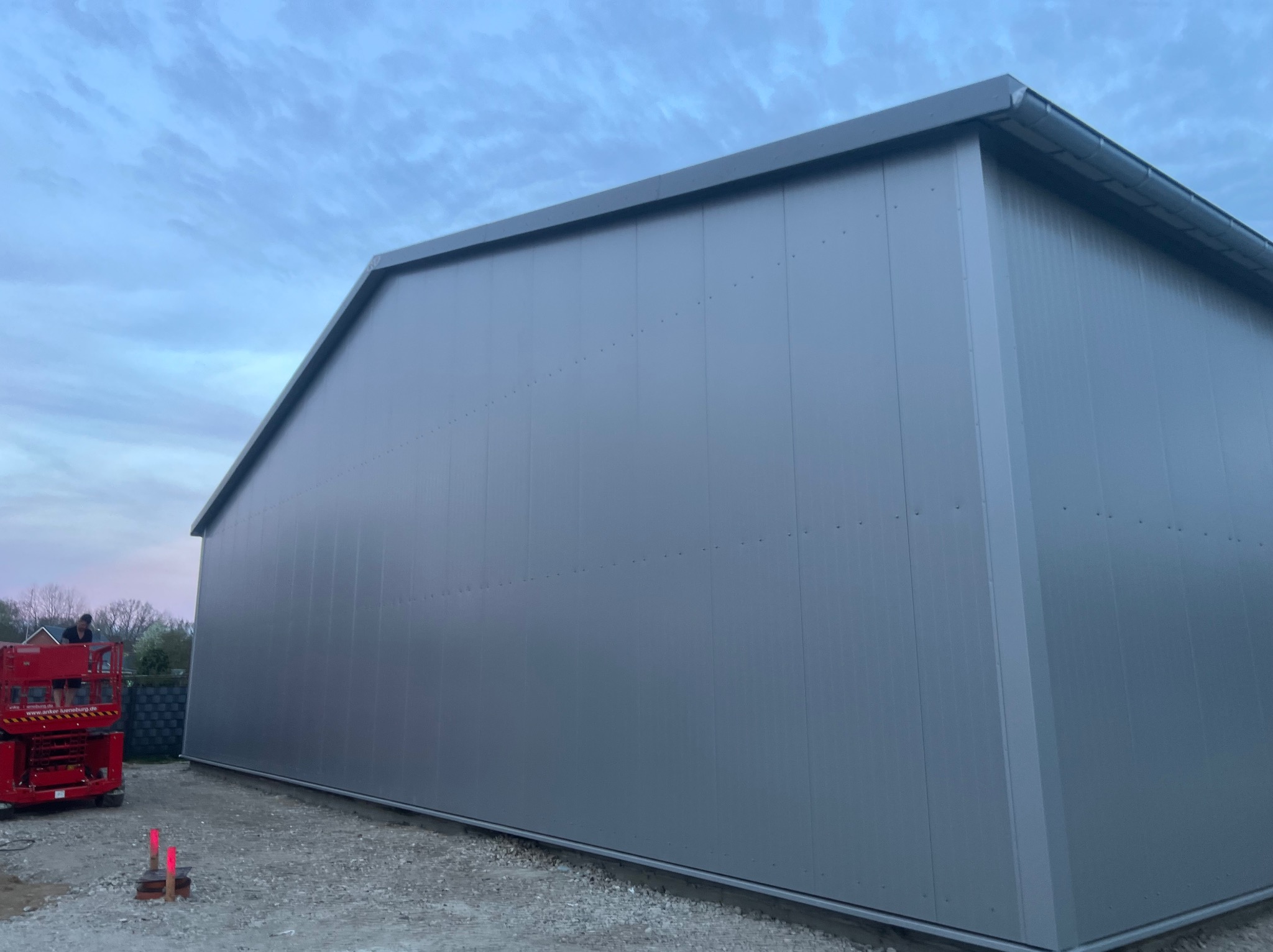 Szara hala produkcyjna z płyt warstwowych, widoczny podnośnik koszowy z pracownikiem na tle zachmurzonego nieba.