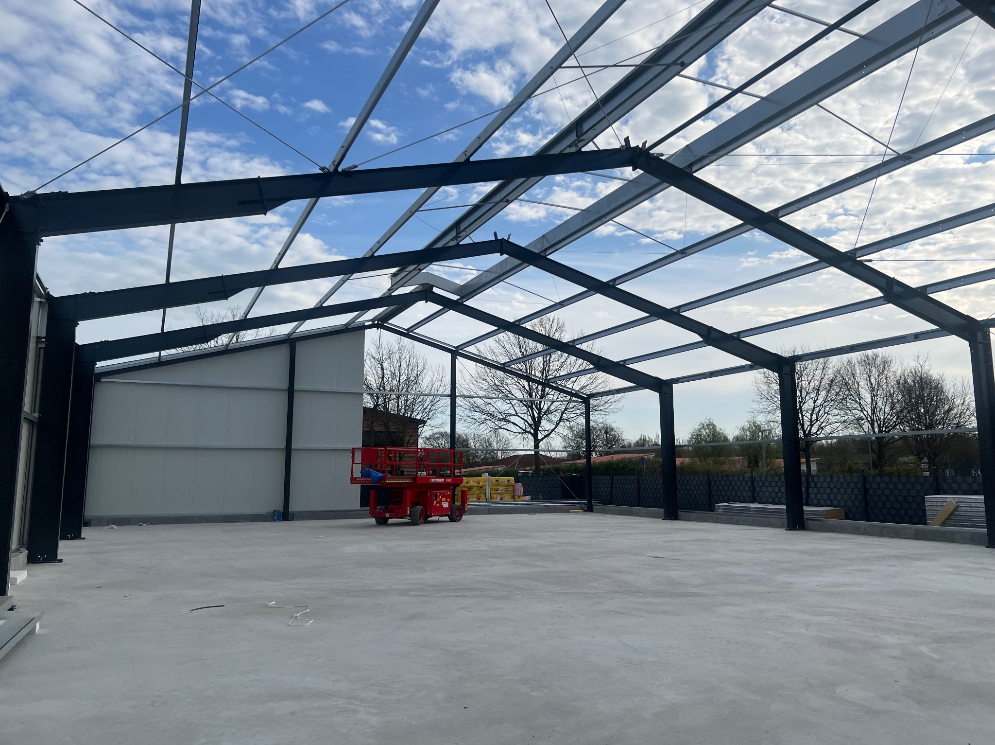 Konstrukcja stalowa hali w trakcie montażu, widoczny szkielet dachu i ścian, podnośnik koszowy wewnątrz, płyty warstwowe przygotowane do montażu, błękitne niebo w tle.