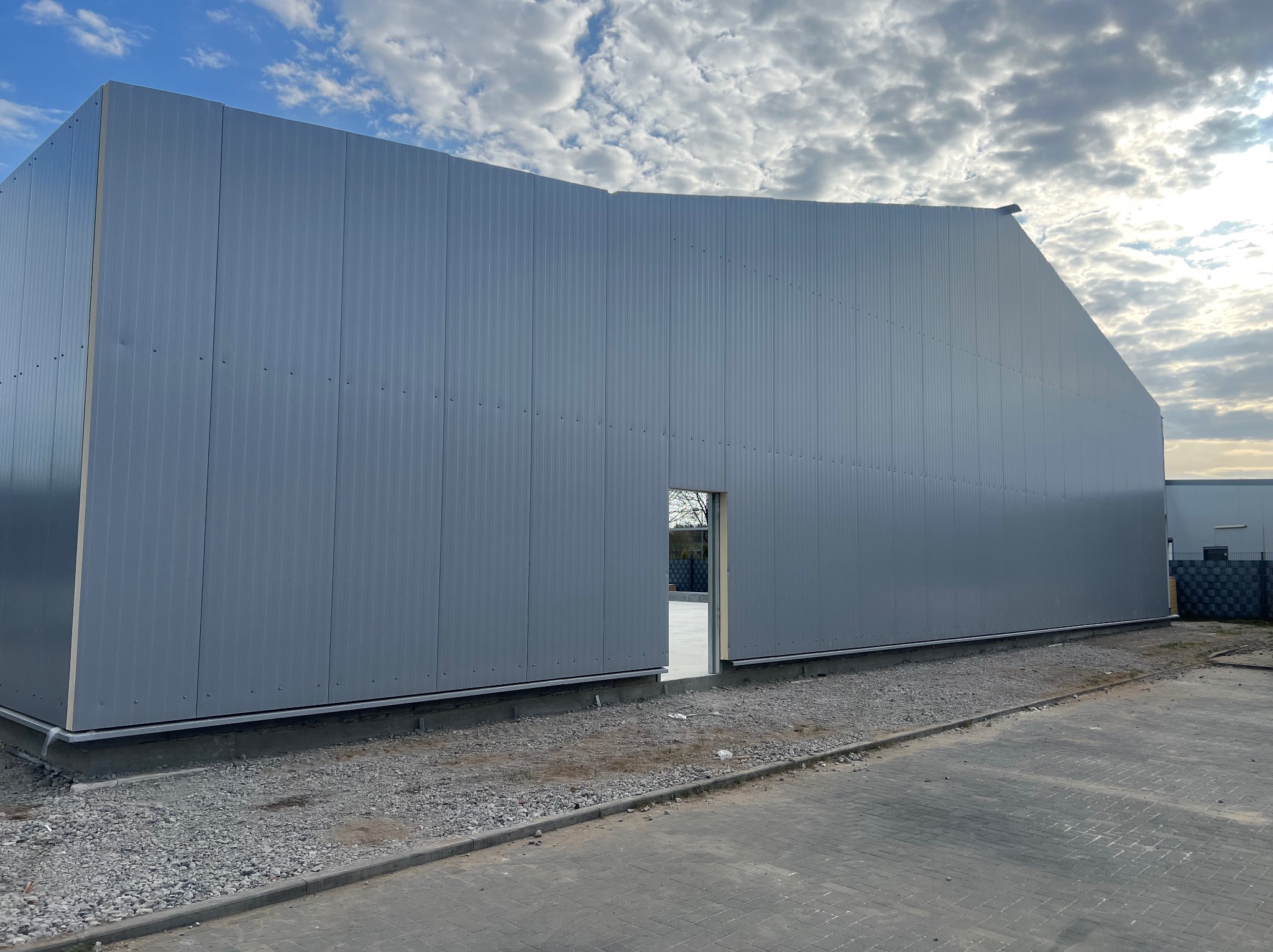 Budynek w trakcie montażu z płyt warstwowych, widoczne panele ścienne i otwór drzwiowy, na zewnątrz plac budowy.