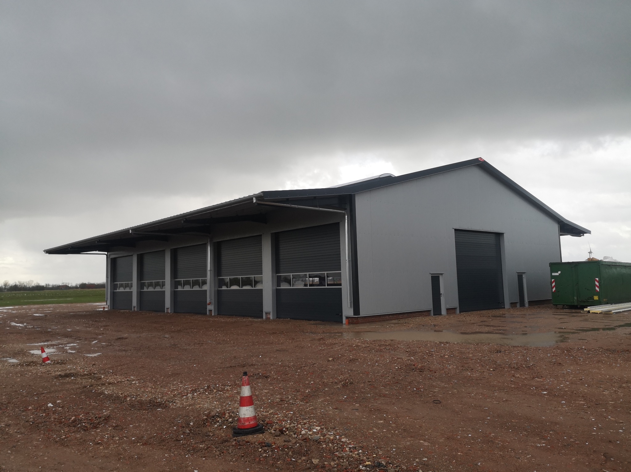 Nowoczesny, szary budynek przemysłowy z czterema bramami rolowanymi i płaskim dachem, widoczny na terenie budowy w pochmurny dzień, z zielonym kontenerem w tle.