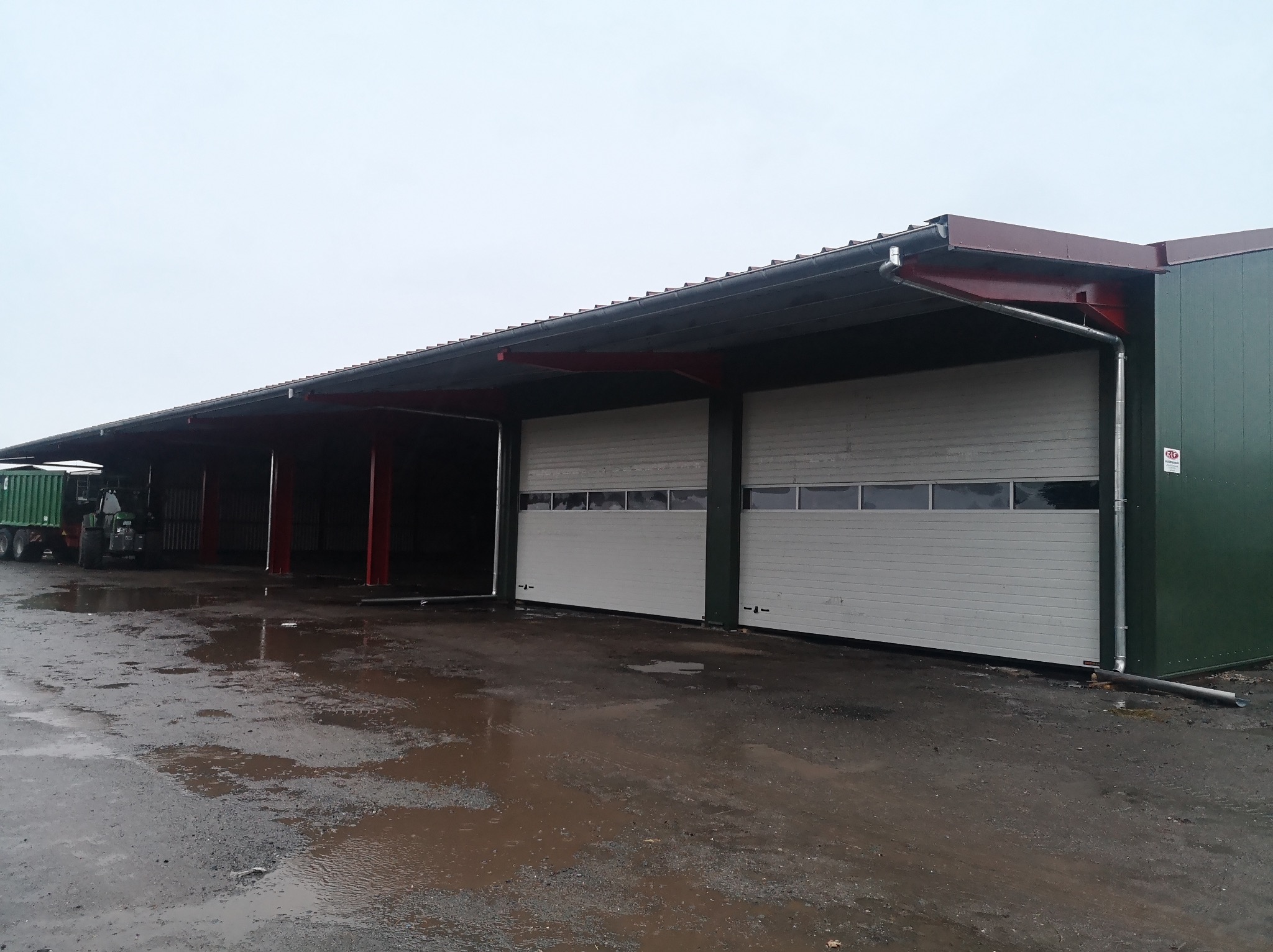 Zadaszona wiata z konstrukcją stalową, blachą trapezową i bramami segmentowymi, widoczny traktor z przyczepą w tle, teren po deszczu.