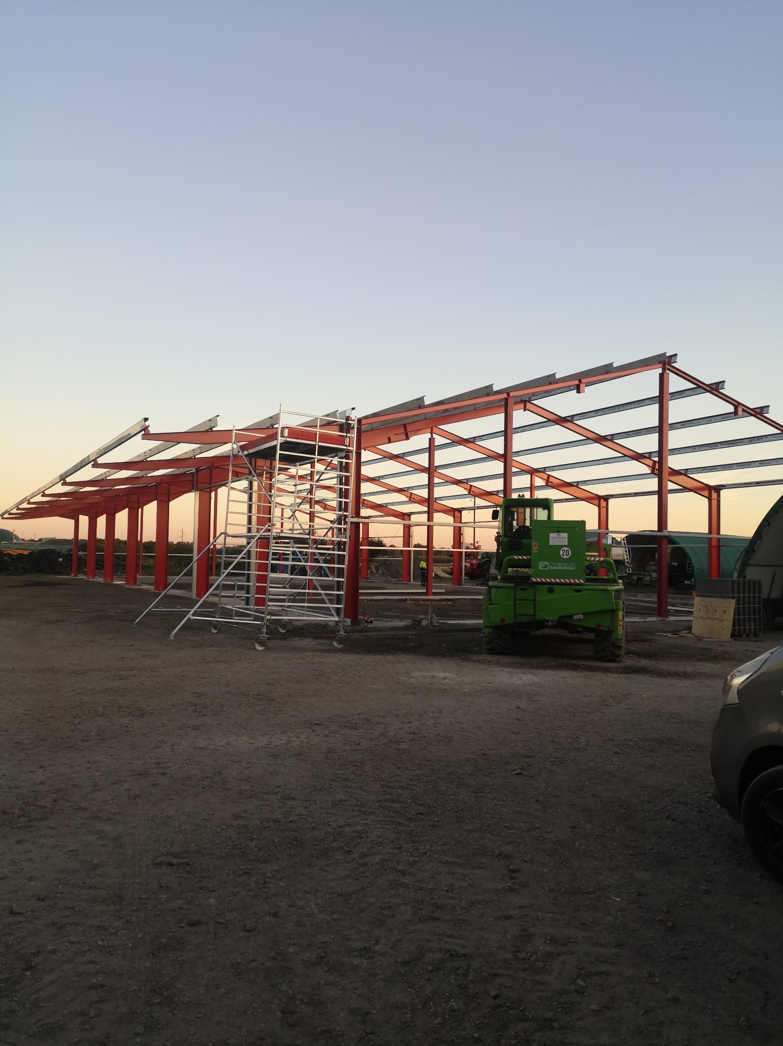 Konstrukcja stalowa budynku w trakcie montażu, czerwone słupy i belki, na placu budowy podnośnik i rusztowanie, zachodzące słońce w tle.